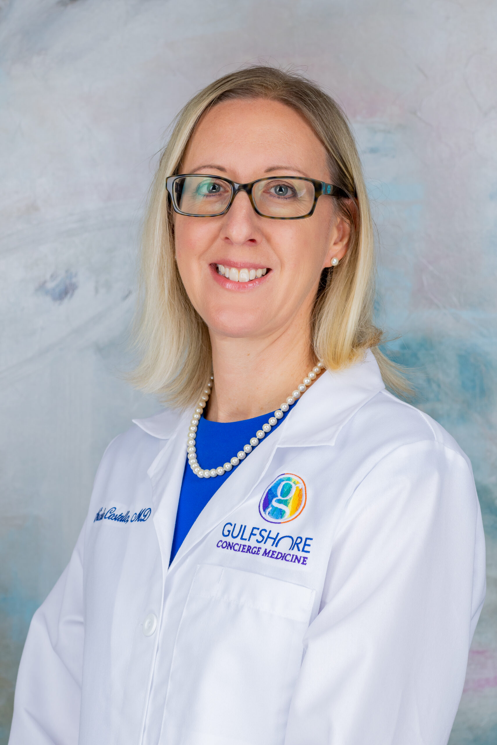 Gulfshore_Concierge_Medicine_2024_Dr._Nicole_Costello_Headshot_@ ES9_Photography_2024 A female doctor wearing glasses and a white coat smiling.